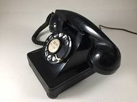 Side view of black rotary dial telephone showing coiled handset cord, rotary dial with finger stop and base with feet.