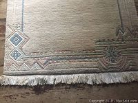 Overall view of corner and part of carpet showing wool material, cream background and colored geometric patterned border with fringed edge.