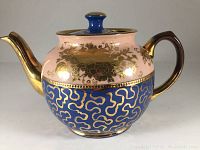 Front side of the teapot showing blue with gold abstract pattern and pink gold floral design, showing handle and spout
