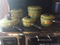 Cookware set displayed on stove showing olive green pieces decorated with colorful flower border and matching lids with black knobs.