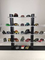 Full view of 25 Hot Wheels cars displayed on a clear acrylic four-level stand showing a range of models and colors arranged by rows.