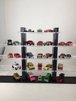 Full view of the 27 toy cars on the clear acrylic display stand showing four levels with various small models in different colors such as red, pink, yellow, orange, blue, and green.