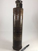 Front view of the vintage metal La France fire extinguisher with embossed branding and instructions, includes pen for scale.