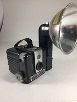 Side and front view of Kodak Brownie Hawkeye Flash Model camera showing lens, viewfinder, and attached flash reflector bowl