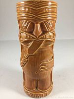 Front view of vintage brown ceramic tiki mug featuring a stylized figure licking a paddle, with detailed facial features.
