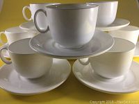 Eight white bone china cups each paired with a saucer, arranged in a cluster on a yellow surface.