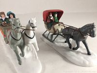 Side view of two Dickens Village ceramic sleighs with horses and figures on white snow-like bases, close view of details and paint.