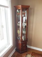 Front view of the wood and glass curio cabinet positioned near a window, showing the upper glass door section and lower wood storage door.