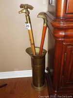 Three wooden canes standing inside a tall brass holder against a wall and a wooden cabinet.
