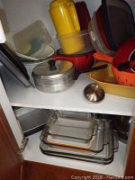 Shelf with assorted plastic storage containers, metal saucepan with lid, yellow ceramic baking dish, orange Le Creuset Dutch oven lid, and glass baking dishes.