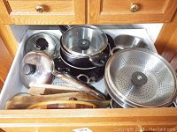 Drawer with stainless steel pots and pans, multiple glass and metal lids with black knobs, and wooden cutting boards lying flat.