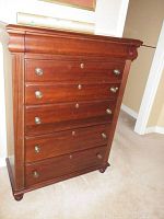 Front view of tall wooden dresser showing four drawers with keyholes and metal knobs, decorative molding on top, and rounded feet at bottom