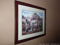 Framed print hung on wall showing the village scenery with water reflection in a reddish wooden frame.