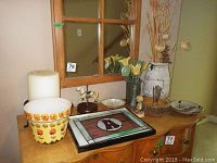 Photo showing a tabletop with various decorative items including floral ceramic pot, candle, stained glass panel, and dishes.