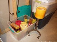 Assorted used candles in a plastic container, some pillar and block candles, alongside a metal tripod candle holder with a yellow pillar candle on top