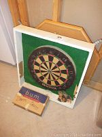 Dartboard mounted in a white wooden frame with a green felt backing, box of darts included below