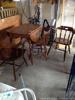 Overall view of colonial style wooden drop-leaf table and three chairs on concrete floor showing condition and style.