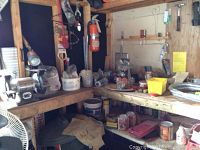 Photo of a workshop corner with a workbench, various containers holding nails, bolts, and other small parts, paint cans, and other assorted items. Fire extinguisher mounted on wall is visible but not included in the lot.