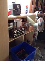 Shelf with assorted items including small drawers, buckets, boxes, a standing fan, and a blue recycling bin on the floor.