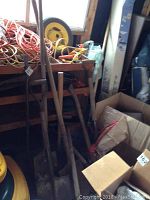 Photo showing long-handled shovels, rakes and other yard tools leaning against a wooden surface inside a shed.