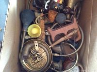 Top view of a cardboard box containing multiple vintage bulb horns with rubber squeeze bulbs, brass wall plaques with classical scenes, and other metal decorative pieces.