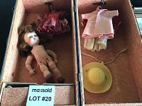 Nude vintage doll with loose arms laying inside pink fabric-lined box along with several doll dresses and a yellow straw hat.