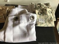 Photo showing folded white curtain panels with metal grommets and patterned green panels partially visible behind.