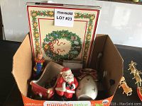 Box containing a Fitz and Floyd sleigh vase with Santa Claus figure, Christmas placemats with wreath design, and other holiday figurines and ornaments.