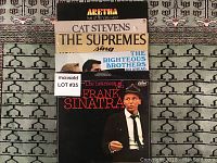 Photo shows six LP vinyl record albums stacked vertically on a patterned carpet. Visible album covers include Aretha Franklin, Cat Stevens, The Supremes, The Righteous Brothers, and Frank Sinatra.