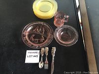 Image showing the full lot arrangement on black surface including amber glass plates stacked, a large amber child's plate with etched designs, a pink glass bowl, a pink glass mug, and the set of stainless steel flatware with bear handles.