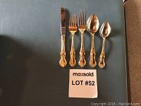 Gold plated flatware displayed individually: knife, two forks, two spoons with ornate handles.