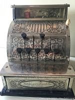 Front view of toy metal cash register showing decorative engraving, six black lever buttons, and Benjamin Franklin label plaque.