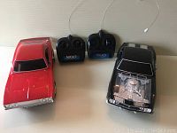 Side-by-side top view of red Camaro and black Charger Nikko remote control cars with corresponding black remote controllers placed between them showing antennas and buttons.