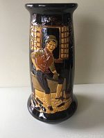 Front view of a tall vintage vase with a glossy dark glaze and raised relief depicting a man sitting outside a building.