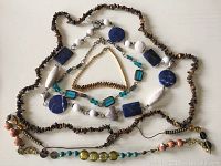 Top down photo showing five necklaces with mixed beads and stones in blue, white, brown and metallic tones laid out on a white surface.
