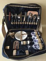 Opened navy blue picnic bag showing organized stainless steel cutlery, white plates, and glasses with fabric covers inside.
