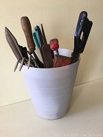 White metal bucket holding various garden hand tools including a hand rake, trowels, and other small gardening tools with wood and plastic handles.