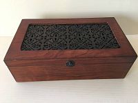 Closed wooden wine bottle box showing smooth finish and black metal decorative top panel with floral geometric cutouts, front metal clap lock visible.