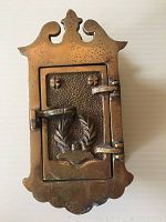 Front view of closed brass door peek box showing ornate crest and embossed motif