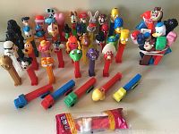 Photo showing a collection of 40 colorful PEZ candy dispensers with various cartoon and pop culture character heads arranged upright, including duplicates and one in original package at the front.