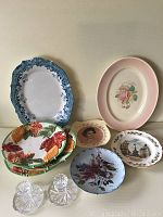 Group photo of six vintage plates with varied floral, portrait, and scenic designs and two clear crystal candle holders with cut geometric patterns.