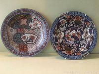 Photo showing two porcelain plates side by side on a shelf, each with intricate decorative patterns in blue, rust, and white colors.
