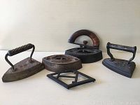 Photo showing three antique sad irons and one trivet stand, including one with a wooden handle.
