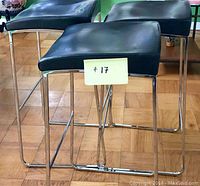 Three contemporary black leather bar stools with chrome metal legs on hardwood flooring.