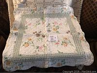 Small square quilt with floral patterns and green border, displayed on a chair with a crocheted cover behind it.