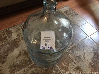 Photo of large clear glass jug on tiled floor showing the jug's overall shape and size relative to the tiles around it.