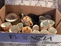 Box showing assorted small tea set pieces with floral teapot, cups, saucers, creamer, sugar bowl, and black creamer pitcher.