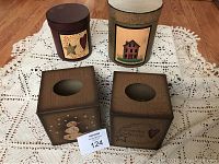 Four items on crocheted white tablecloth: two square tissue boxes with oval or rounded openings, and two round containers with country house artwork.