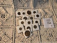 14 assorted tokens and coins arranged on a crocheted blanket, each secured in a white cardboard holder with clear window, visible numbers and designs on some tokens.