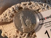 Front view of silver-colored collectible coin featuring Statue of Liberty and LIBERTY inscription in a clear plastic protective case, resting on a crocheted surface.